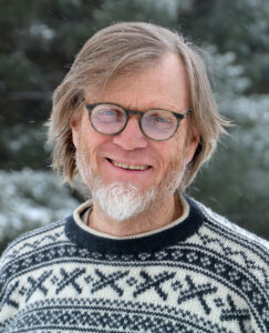 A man with a beard, wearing a sweater and glasses, smiles to camera.