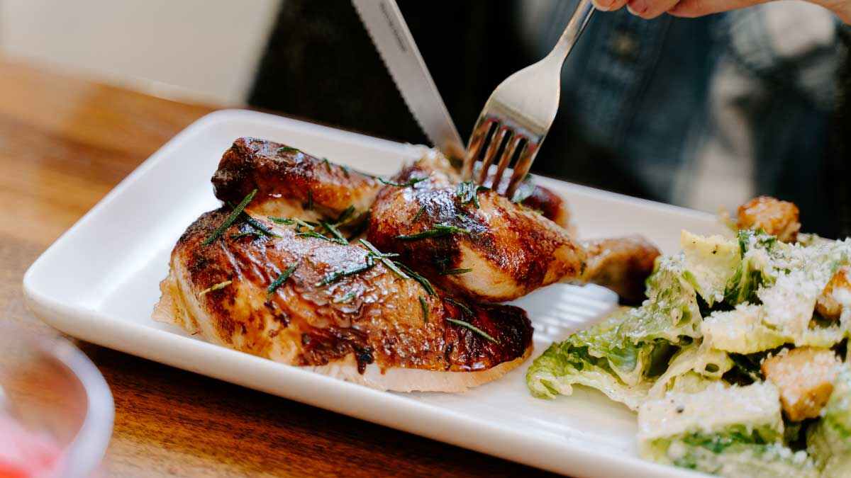 A fork and knife cutting into cooked chicken alongside a salad on a white plate
