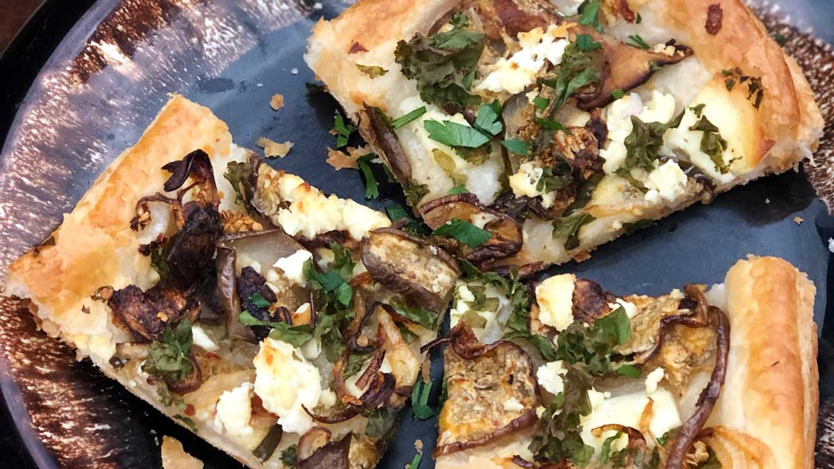 Small pieces of flatbread with eggplant slices, onion slices, small leafy greens and small cheese crumbles
