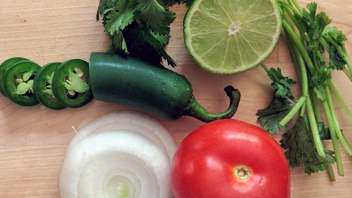 Green jalepeno slices, greed leaves, a lime slice, sliced onion and a red tomato on a wooden surface