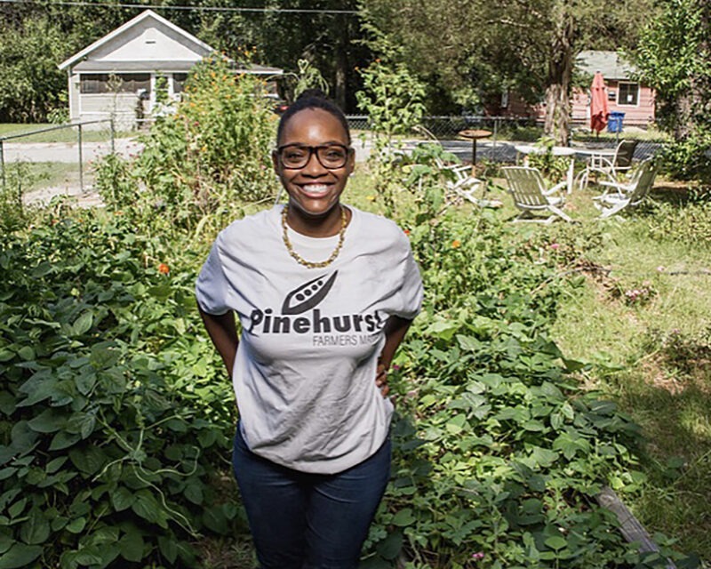 A woman in a grey t-shirt stands proudly smiling to camera in front of a small garden.
