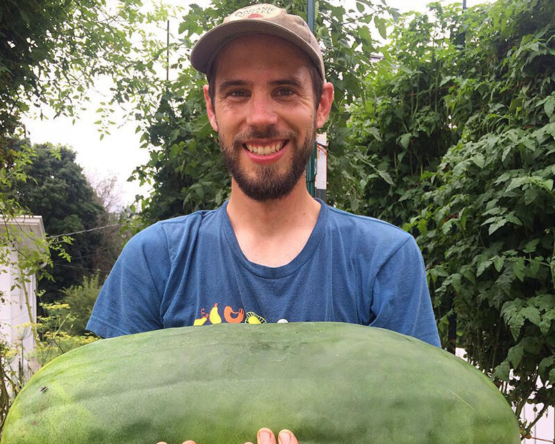 A man in a baseball cap smiling and holding a large oval watermelon