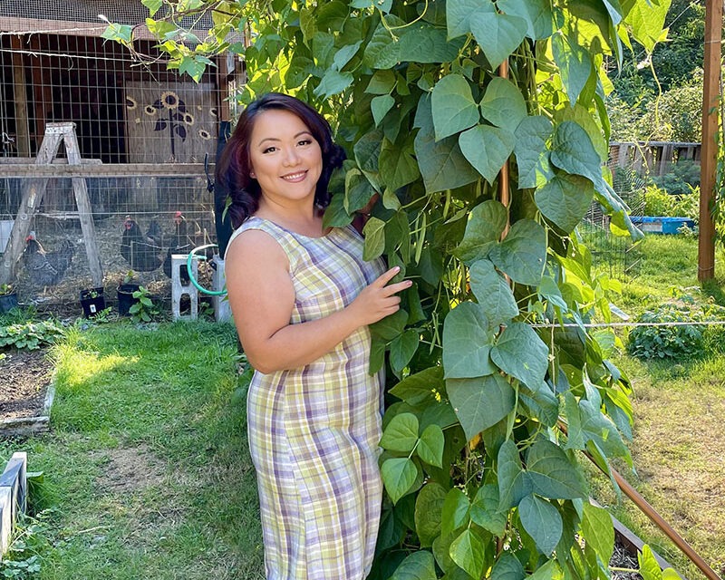 A woman in a plaid dress smiles and poses next to a tall leafy plant