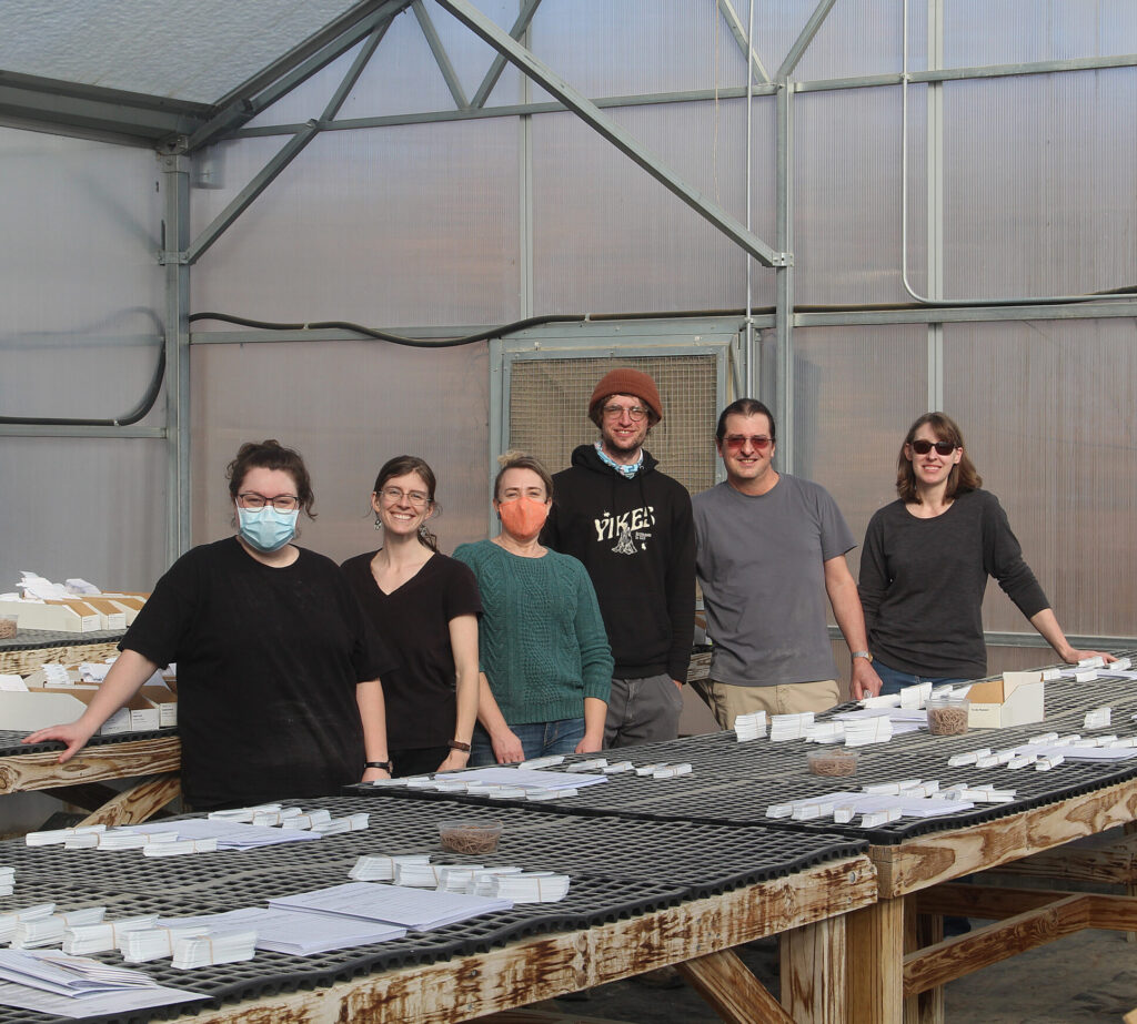 A group of people smile to camera in a green house with seed packets organized on the tables.