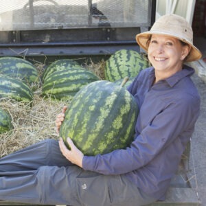 A woman with a wide brimmed hat sits with a large watermelon in her lap with watermelon behind her.
