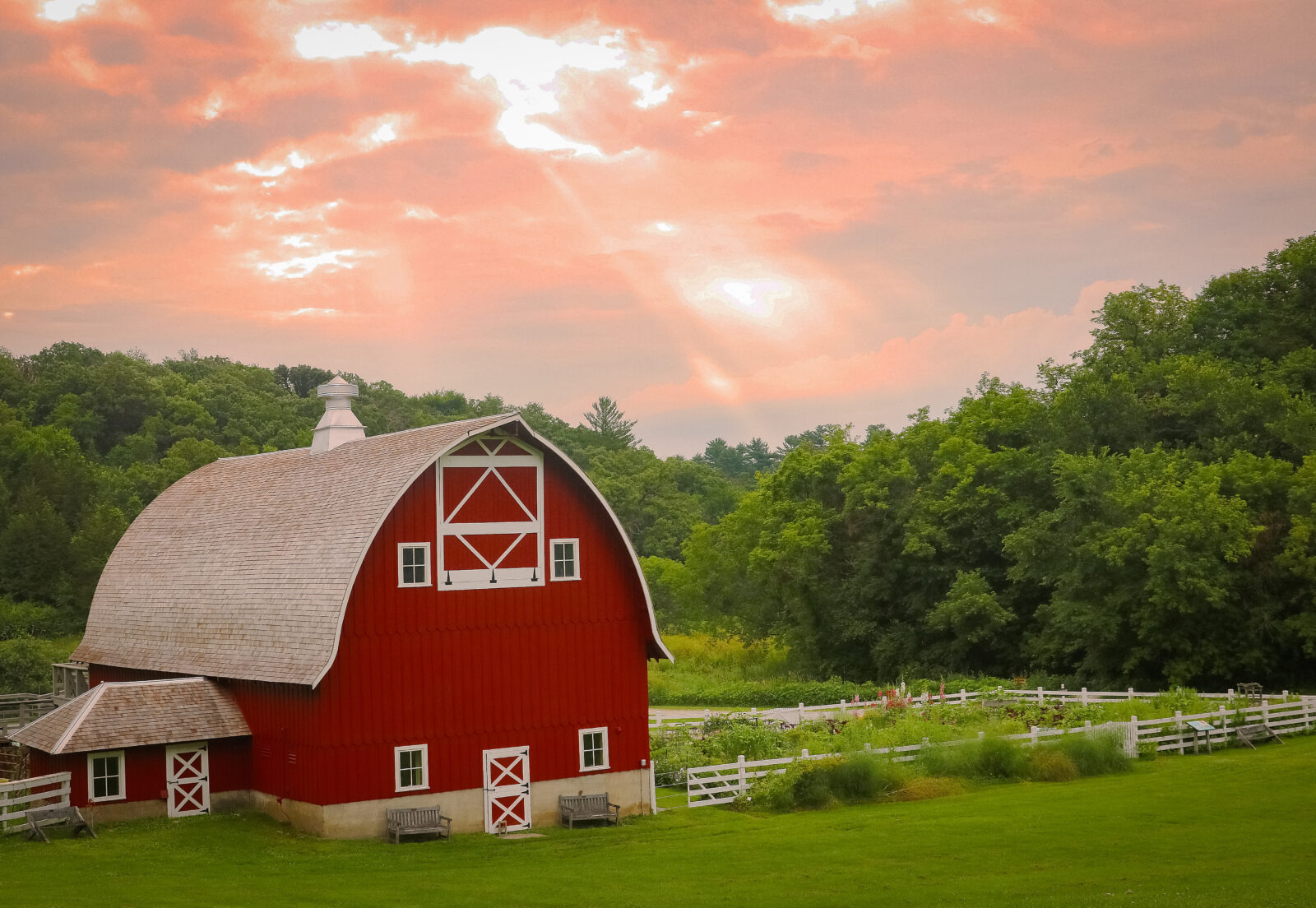 A red barn next to trees and a garden with a pink sunset in the sky