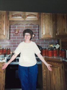 A woman stands in a kitchen next posed in front of many jars of tomato juice cocktail on the counter.