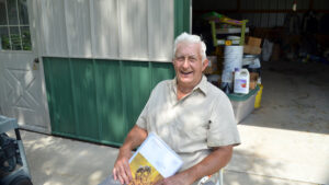 A man in a tan shirt sits holding a magazine in front of a large garage