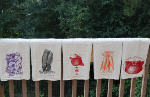 Five flour sack towels with drawings of different plants hanging on a railing outdoors