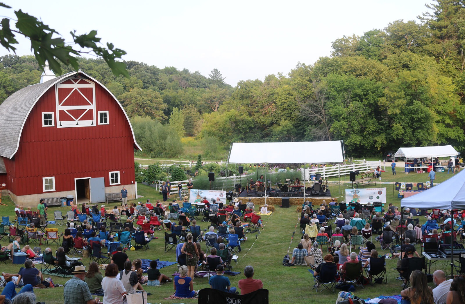 Many people gathered outside in chairs facing a stage next to a red barn