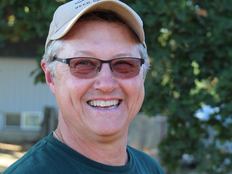 A man in sunglasses and a baseball cap smiles outdoors