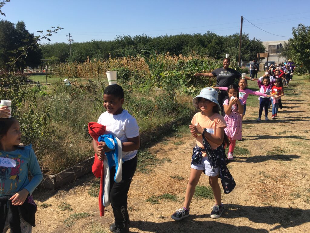 Young students and their teacher walk through a garden while balancing cups on their heads.