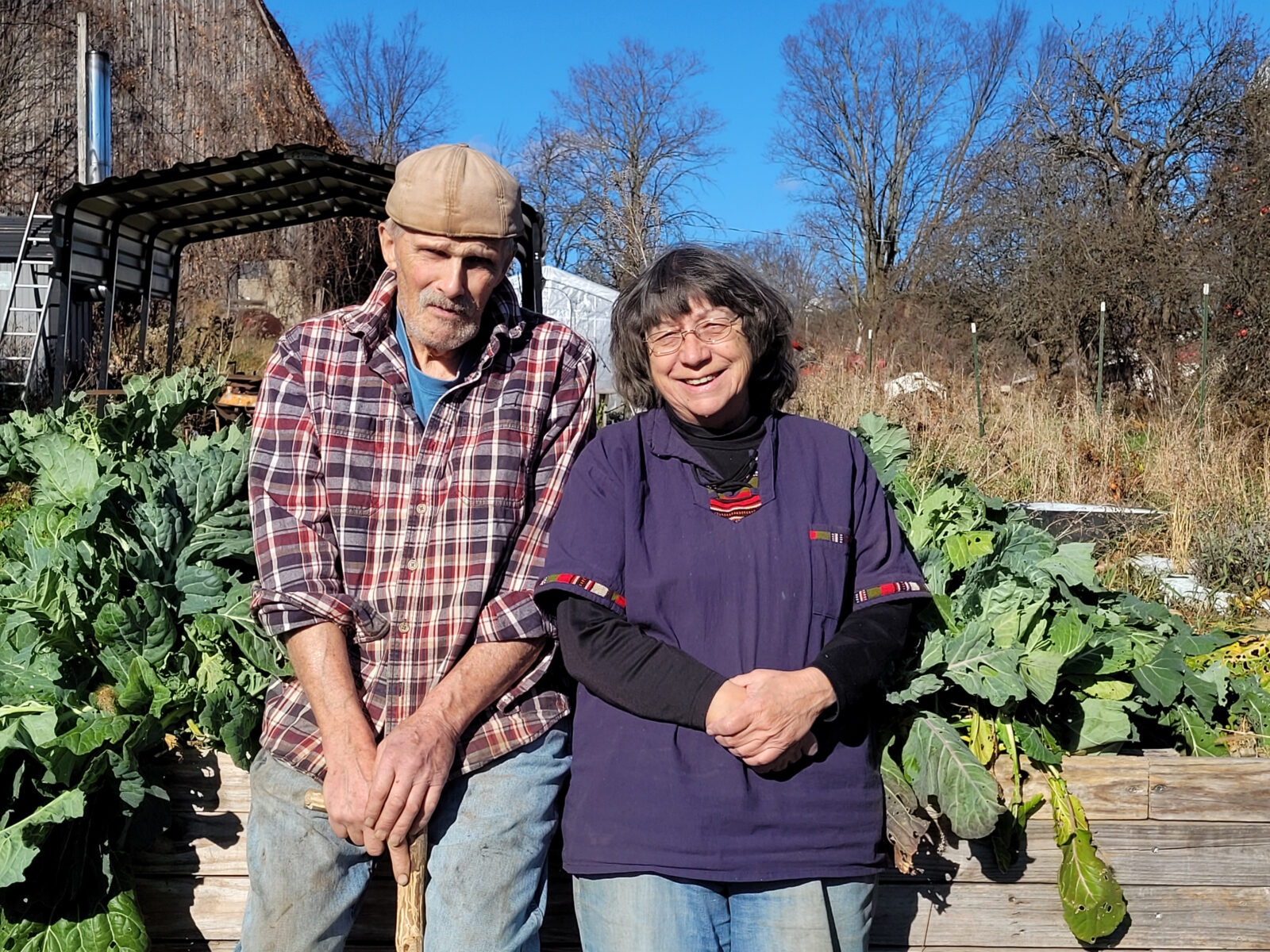 man in plaid shirt and woman in purple sweater stand in front of green garden