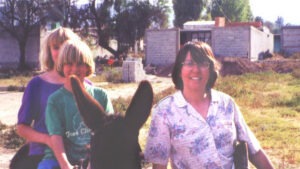 A woman and boy sit on a donkey next to another woman standing, in front of several brick building structures