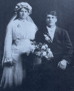 A man sits next to a woman standing in wedding attire, with a basket of flowers between them.