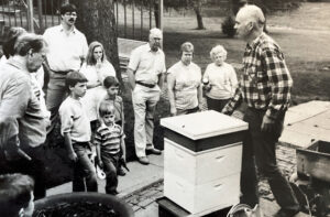 A black and white photo of a man conducting a bee keeping demonstration to a group of adults and children.