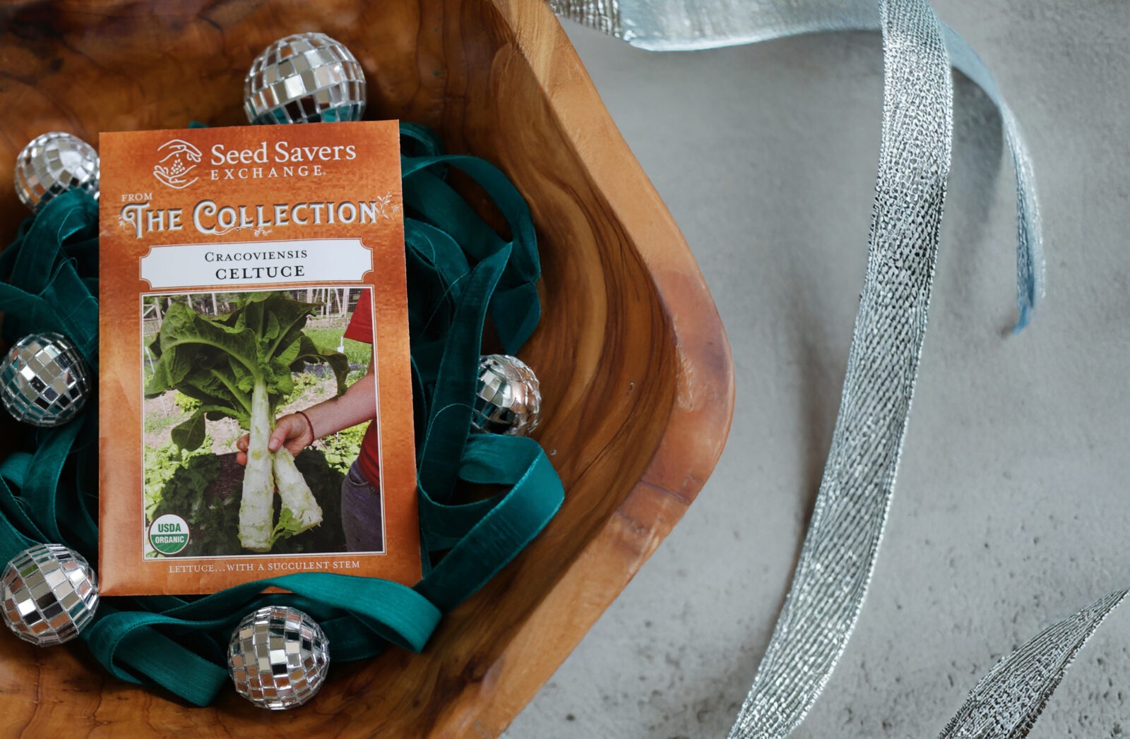A 'Cracoviensis' celtuce seed packet with some green ribbon and small disco balls in a wood bowl next to some silver ribbon