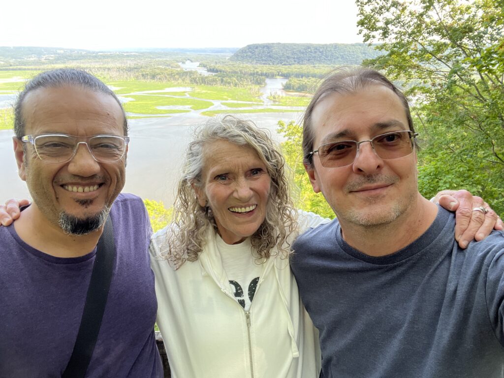Two men and a woman smile with their arms around each other with a beautiful valley in the background