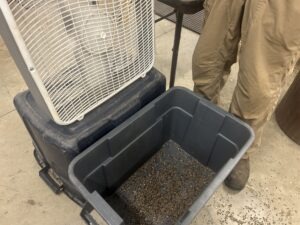 A box fan on top of an overturned bucket with a bucket of seeds in front of it