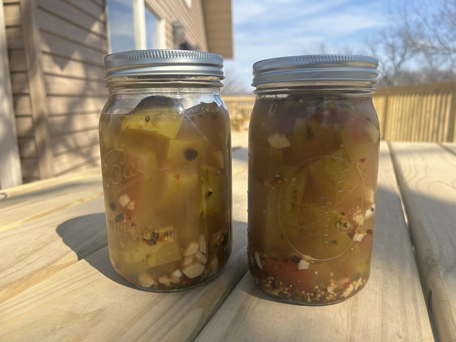 Two large jars filled with pickled watermelon rinds along with spices and chopped garlic sit outdoors on a picnic table
