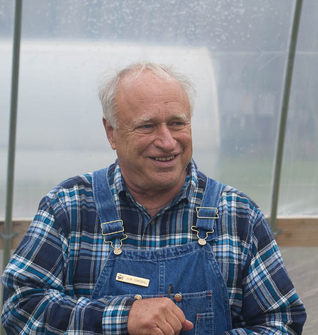 A man in overalls smiles at another man in a greenhouse