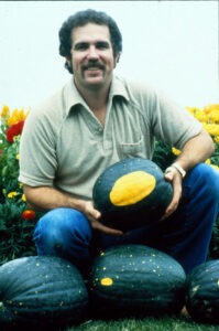 A man poses holding a dark green 'Moon & Stars (Van Doren)' watermelon with a huge yellow blotch, with more watermelons around him.