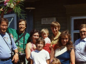 A group of six adults and two children stand and smile in front of a house