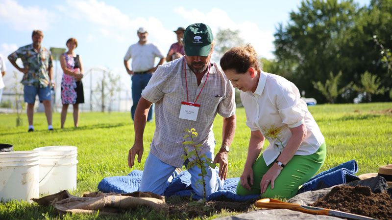 Two people kneel in front of a newly planted tree with more people watching on in the background.