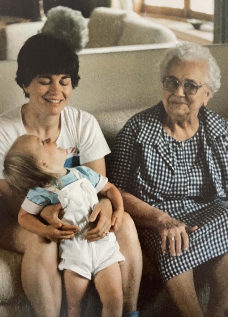 Two women sit on a couch. The younger woman is holding a young child who is looking up at her.