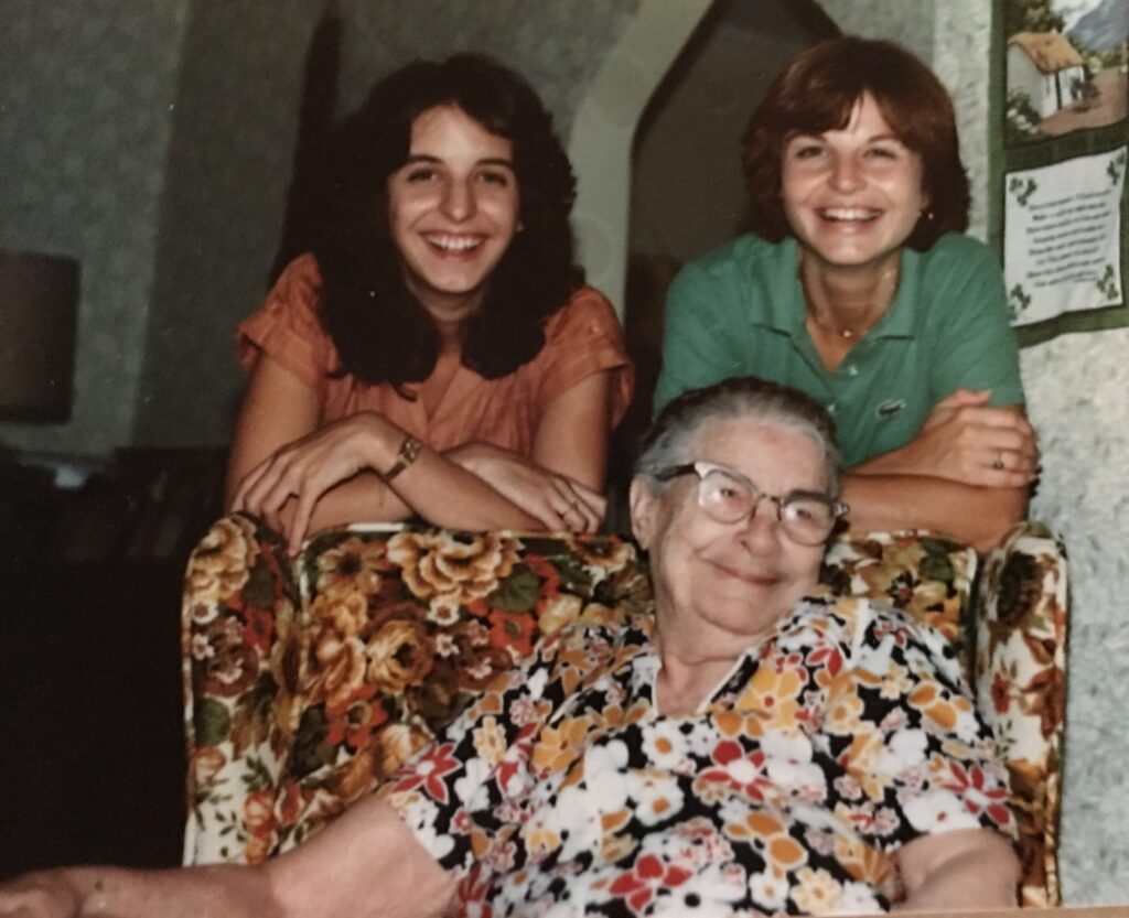 An older woman sits in a floral armchair with two younger women leaning on the chair behind her.