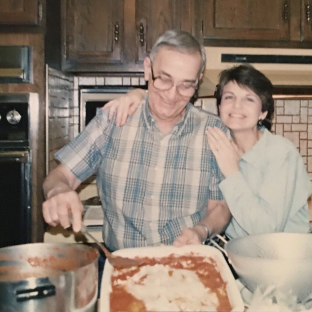 A man scoops cheesy pasta from a baking pan in a kitchen with a woman standing next to him and holding his shoulders.