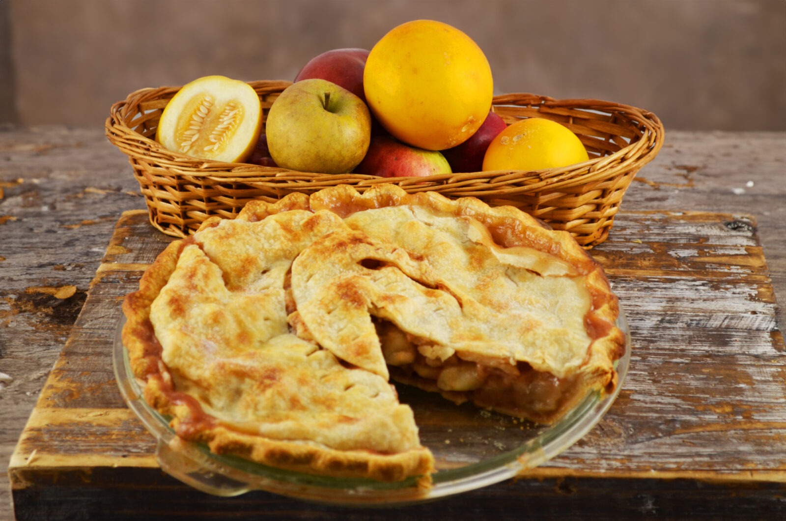 A pie with a slice taken out on a wooden table, and a basket of small yellow melons and apples.
