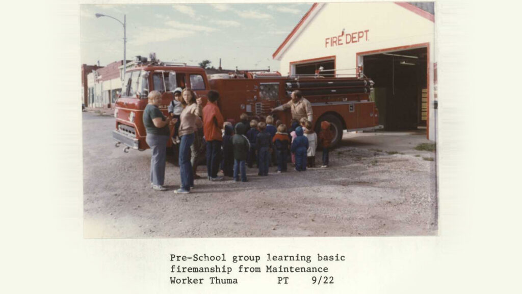 A group of adults and children gather next to a firetruck outside of a firehouse garage with text reading "Pre-School group learning basic firemanship from Maintenance Worker Thuma. PT 9/22"