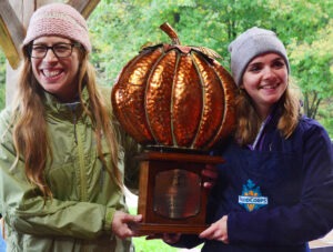 two women wearing stocking hats and coats hold a large, bronze pumpkin trophy