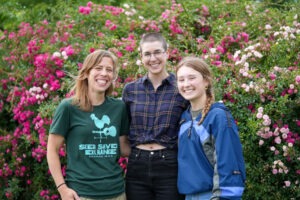 Three women stand in front of a backdrop of flowers.