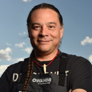 A man smiles to camera with blue sky and clouds behind him.