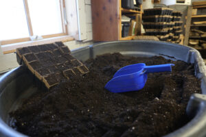 A block of small cell compartments filled with soil, resting in a tub of soil and a blue scooper.