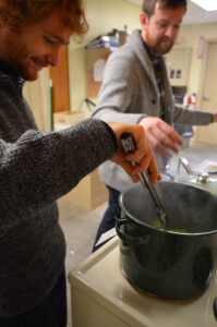 A person stirs a pot while another person prepares food in the background