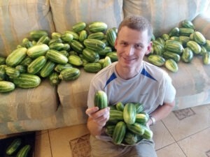 man in grey T-shirt, holding green cucumbers, sits in front of couch with dozens of green cucumbers