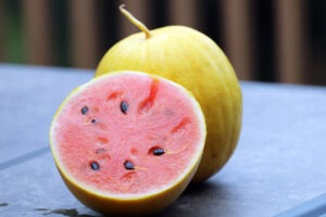 A small, round, yellow watermelon and a halved yellow watermelon with pink flesh sit on a table