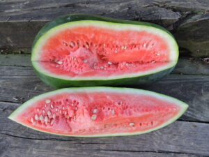 A long watermelon with a slice taken out lengthwise lies on a wood table, exposing bright pink flesh