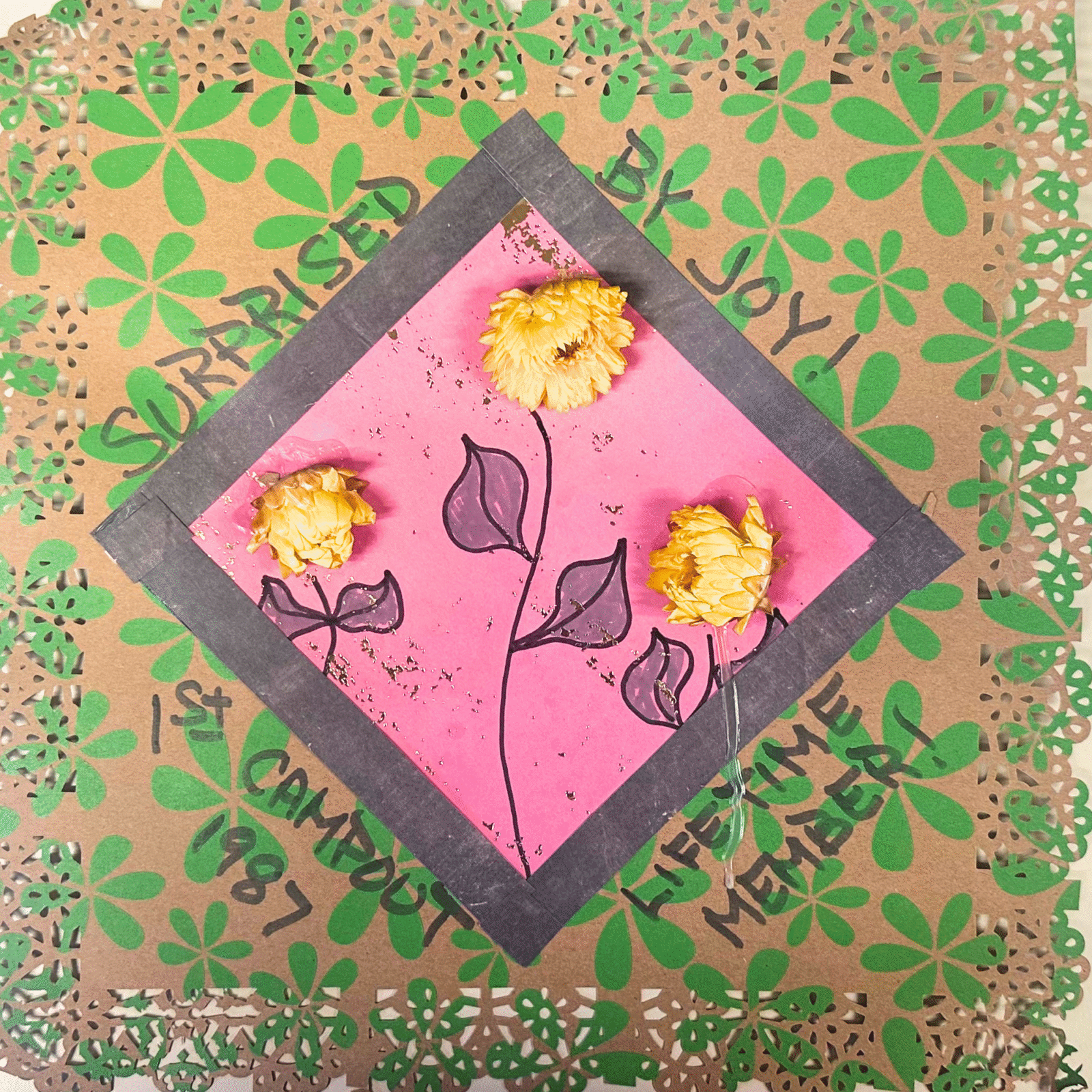 A brown scrapbook page with green flowers and a pink square with yellow flowers