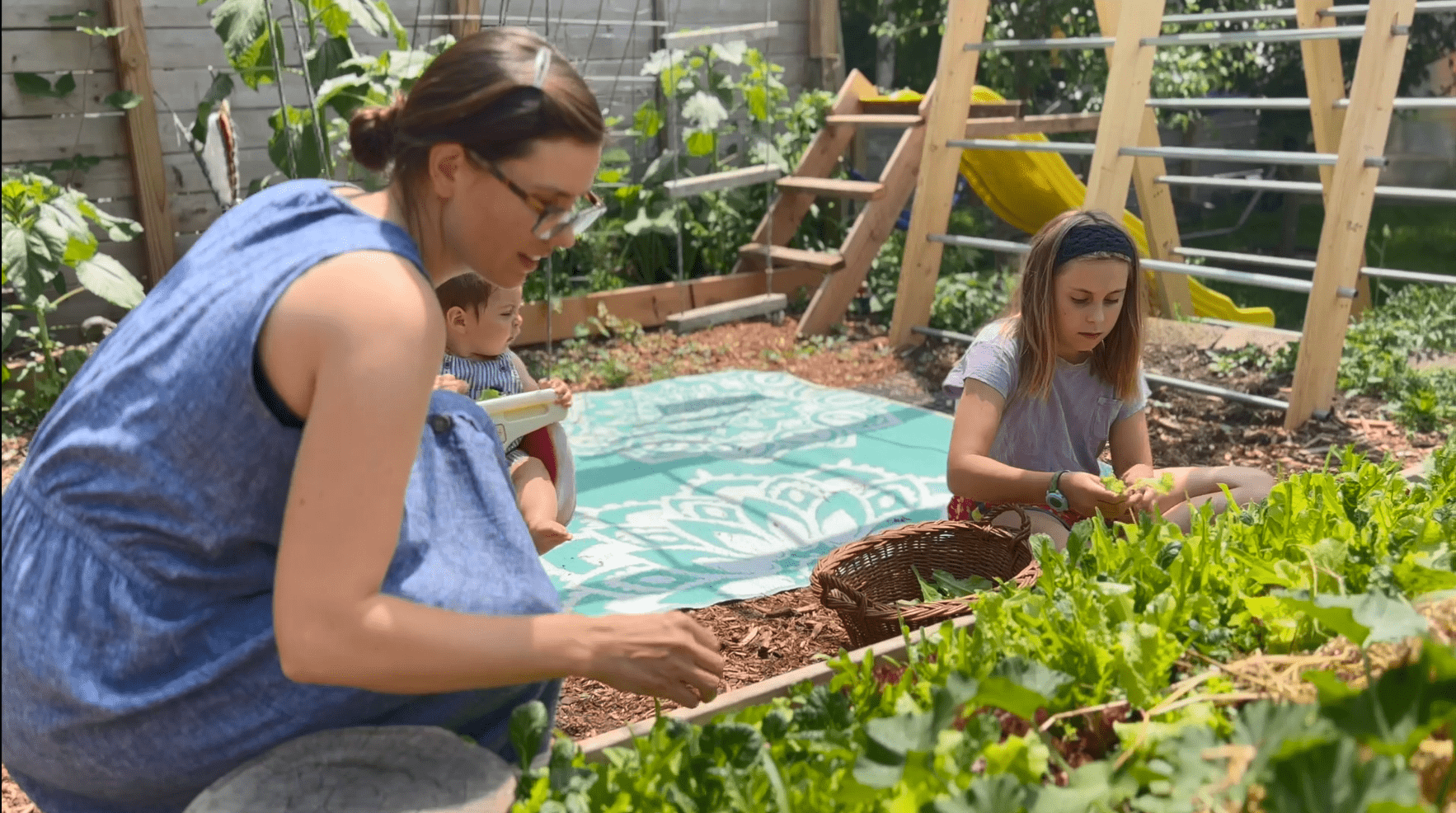 A woman and a young girl kneel next to a garden bed outdoors, picking vegetables. An infant sits in a swing behind them.