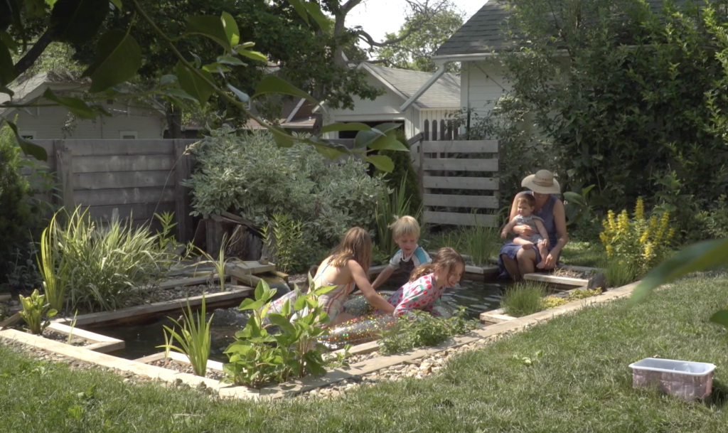 Several children playing in a small outdoor pool in a backyard while a woman holding a baby sits with her feet in the pool