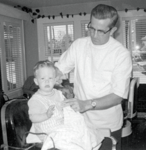 A baby sits in a high chair next to a man cutting the baby's hair.