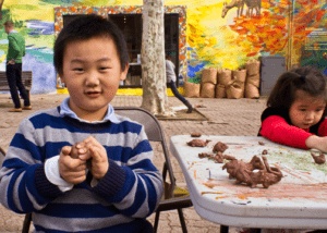 A young boy poses holding clay next to a folding table with various small clay structures on it