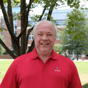 A man in a red polo shirt, with a tree behind him, smiles to camera.