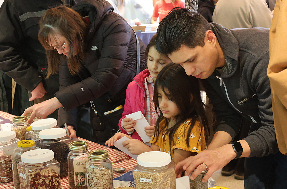 Crowd closely inspects a table of seeds in various containers.