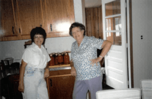 Two women pose standing in a kitchen in front of many jars of canned tomatoes on the counter.