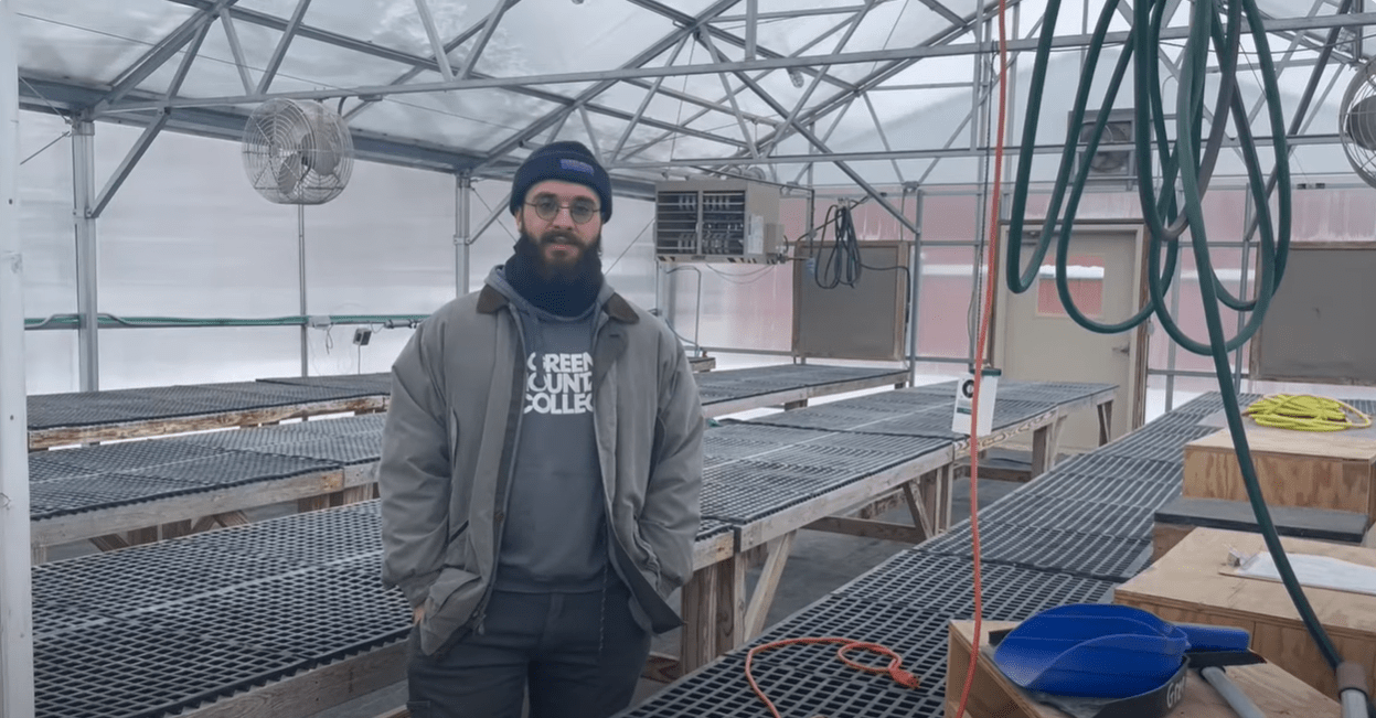 A man wearing a coat and hat stands in a greenhouse